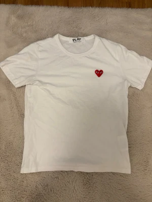 Vit Comme des Garçons PLAY t-shirt - Vit t-shirt från Comme des Garçons PLAY med det ikoniska röda hjärtat med ögon broderat på bröstet. Klassisk rund halsringning och korta ärmar. Tillverkad i mjuk bomull för en skön och avslappnad känsla. Passar dig som är ca 167-174 cm