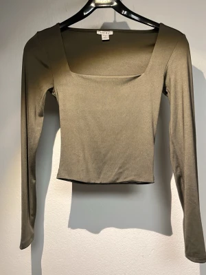 Olivgrön långärmad topp från Amisu - Snygg olivgrön långärmad topp från Amisu med fyrkantig ringning och croppad passform. Tillverkad i mjukt stretchigt material som sitter skönt på kroppen. Perfekt att styla med höga jeans eller kjol för en trendig look.