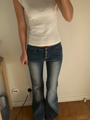 Blå bootcut jeans med knappar - Säljer ett par blå bootcut jeans med låg midja och fyra synliga knappar framtill. Jeansen har en snygg tvätt med ljusare partier på låren och lätt utsvängda ben. Perfekta för en avslappnad men trendig look.