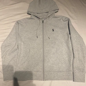 Grå hoodie från Polo Ralph Lauren - Snygg ljusgrå hoodie från Polo Ralph Lauren med dragkedja framtill och klassisk huva. Tröjan har två fickor och den ikoniska lilla loggan broderad på bröstet. Tillverkad i mjukt bomullsmaterial som känns skönt mot huden. Perfekt för en avslappnad look.