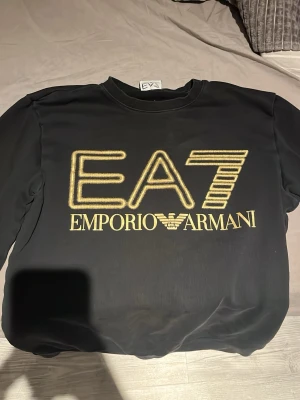 Svart EA7 Emporio Armani hoodie - Snygg svart hoodie från EA7 Emporio Armani med stort guldfärgat tryck på bröstet. Klassisk passform med rund halsringning och långa ärmar. Perfekt för dig som vill ha en stilren och trendig look med exklusiv känsla.