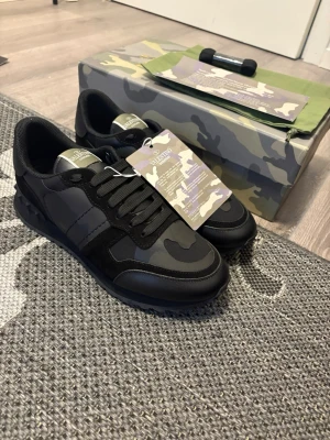 Valentino Garavani , KAN GÅ NER PRIS!!  - Snygga sneakers från Valentino Garavani i svart och mörkgrått med camomönster. Skorna har detaljer i mocka och läder, samt nitar på hälen för en edgy look. Klassisk låg modell med svarta skosnören och robust sula. Perfekta för dig som gillar streetstyle.