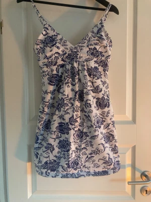 Vit och blå blommig sommarklänning - Söt kortklänning med smala axelband och v-ringning. Klänningen är vit med blått blommönster och har en lätt utsvängd passform. Perfekt för varma dagar och har en mjuk bomullskänsla.