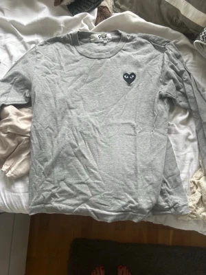 Grå långärmad t-shirt från Comme des Garçons - Säljer en grå långärmad t-shirt från Comme des Garçons PLAY med det ikoniska svarta hjärta-trycket på bröstet. Tillverkad i mjuk bomull och har en klassisk passform med rund hals. Perfekt för en clean och stilren look.