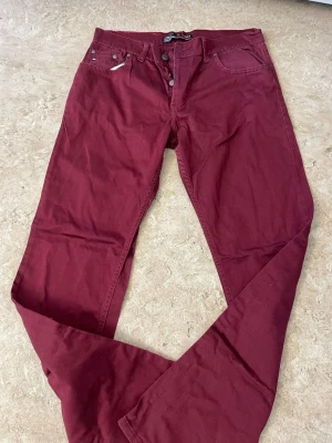 Vinröda raka jeans - Snygga vinröda jeans med rak passform och klassisk femficksdesign. Jeansen har knappgylf och är tillverkade i ett mjukt bomullsmaterial som känns bekvämt hela dagen. Perfekta för dig som vill ha en färgklick i garderoben.