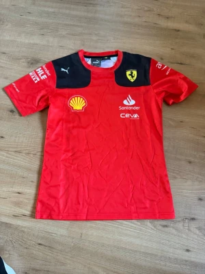 Ferrari F1 t-shirt - Snygg Ferrari F1 t-shirt från Puma i storlek M med svarta axelpartier och flera tryckta loggor som Shell, Santander och Ferrari-emblemet. Perfekt för motorsportfans! Tillverkad i lätt och bekvämt material, kortärmad och med rund hals. 
