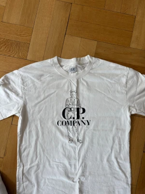 Vit C.P. Company t-shirt med tryck - Säljer en vit t-shirt från C.P. Company med ett stort tryck av en person och loggan på bröstet. Klassisk rund hals och korta ärmar. Perfekt för dig som gillar streetwear och vill ha något stilrent men ändå med en twist.