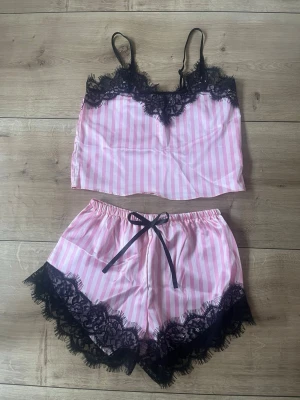 Rosa randig pyjamas med spetsdetaljer - Sött pyjamasset i två delar med rosa och vita ränder. Setet består av ett linne med tunna axelband och svarta spetsdetaljer samt matchande shorts med spetts.  Aldrig använt. 