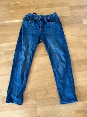 Mörkblå raka jeans med snörning - Säljer ett par mörkblå jeans med raka ben och snörning i midjan. Jeansen har klassiska fem fickor, kontrastsömmar och en knappgylf. Materialet är denim i bomull och passformen är avslappnad. Perfekta för en chill och avslappnad stil.