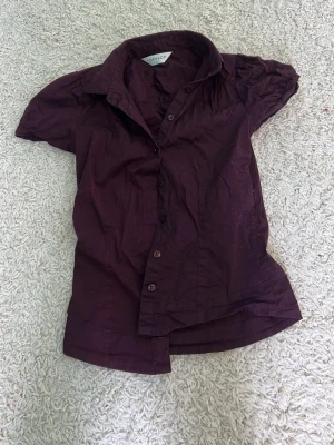 Mörklila skjorta från Topshop - Kortärmad skjorta från Topshop i en snygg mörklila färg. Skjortan har klassisk krage, knappar framtill och lätt puffade ärmar. Tillverkad i bomull som känns skön mot huden. Perfekt för dig som vill ha en enkel men stilren look.