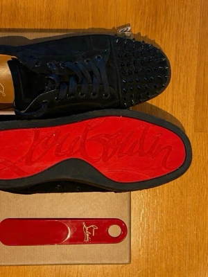 Svarta Louboutin sneakers med nitar - Säljer ett par svarta sneakers från Christian Louboutin i mocka med ikoniska röda sulor och svarta nitar på tån. Skorna har svarta skosnören och en låg silhuett. Perfekta för dig som vill sticka ut med en edgy och lyxig stil.