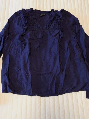 Mörkblå volangblus från Veromoda - Mörkblå blus från Veromoda med långa ärmar och volangdetaljer längs ringningen och framtill. Tillverkad i ett lätt och mjukt tyg som ger en avslappnad känsla. Perfekt för dig som gillar feminina detaljer och enkel stil.