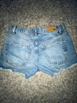Blå denimshorts från 157 - Säljer ett par blå denimshorts från 157 med råa kanter och slitna detaljer framtill. Shortsen har klassisk femficksdesign, bälteshällor och dragkedja. Perfekta för sommaren och ger en avslappnad vibe. Aldirg använt 