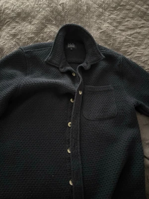 Mörkgrå stickad overshirt  - Snygg mörkgrå overshirt  i grovstickat material. Skjortan har klassisk krage, vita knappar framtill och en bröstficka. Perfekt lager-på-lager-plagg med avslappnad passform och stilren look.