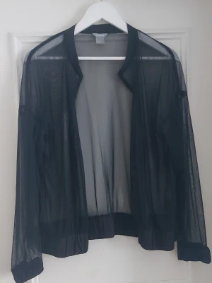 Svart mesh cardigan jacka - Säljer en svart transparent cardigan jacka i mesh med långärmad design och elastiska muddar. Jackan är öppen framtill utan knappar eller dragkedja och har en luftig, modern look som passar perfekt för layering.