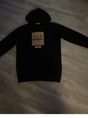 Svart hoodie från Burberry - Snygg svart hoodie från Burberry med stor beige patch och logga på bröstet. Klassisk huva och raka ärmar. Perfekt för dig som vill ha en stilren och exklusiv look. Materialet ser ut att vara mjukt och bekvämt, troligen bomull.
