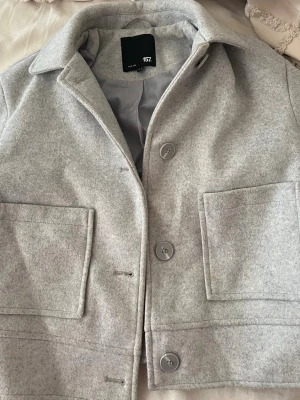 Ljusgrå ullkappa från 157 - Stilren ljusgrå kappa från 157 med rak passform och stora framfickor. Kappan har klassisk krage, tre knappar framtill och är tillverkad i ett mjukt ull-liknande material. Perfekt för dig som gillar minimalistisk och clean stil. Den funkar som m ochså