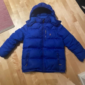 Ralph Lauren jacka blå - Säljer en snygg blå dunjacka från Ralph Lauren med huva och dragkedja. Jackan har en broderad logga på bröstet och praktiska fickor med dragkedjor. Perfekt för kyliga dagar! Är i storlek 14-16 år och passar xxs