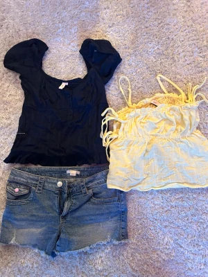 Somrigt klädpaket toppar & shorts - Säljer ett paket med en mörkblå topp med puffärm, en ljusgul croppad topp med knytband och ett par klassiska blå jeansshorts. Perfekt för varma dagar och enkel att matcha med olika stilar. 250kr för allt tillsammans. Tröjorna är storlek 36 och shortsen är xs. Topparna är helt oanvända 