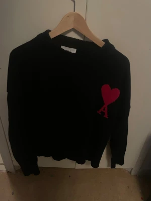 Svart stickad tröja från Ami - Snygg svart stickad tröja från Ami Alexandre Mattiussi med röd hjärtlogga och bokstaven A på bröstet. Tröjan har rund halsringning och ribbade muddar. Perfekt för dig som gillar stilrena plagg med en twist.