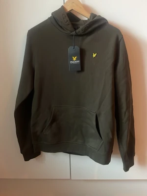 Olivgrön hoodie från Lyle & Scott - Snygg olivgrön hoodie från Lyle & Scott med klassisk gul logga på bröstet. Hoodien har huva med dragsko, stor magficka och ribbade muddar vid ärmar och nederkant. Tillverkad i mjukt bomullsmaterial som är skönt att bära.