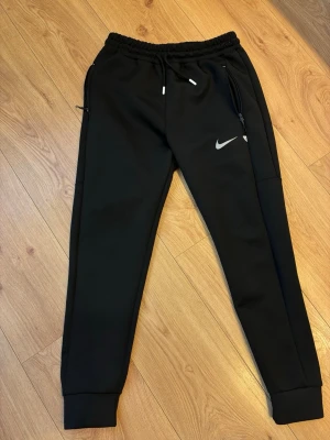  Nike träningsbyxor  - Säljer ett par svarta träningsbyxor från Nike med smal passform och resår i midjan. Byxorna har dragkedjeförsedda fickor, muddar vid bensluten och en vit Nike-logga på vänster lår. Tillverkade i ett mjukt och stretchigt material som passar perfekt till träning.