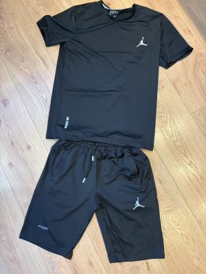 Jordan tracksuit herr - Snyggt svart träningsset från Jordan med t-shirt och shorts. Setet har den klassiska Jumpman-loggan i vitt på både tröjan och shortsen. Tillverkat i ett lätt och ventilerande material som passar perfekt för träning eller sportaktiviteter.