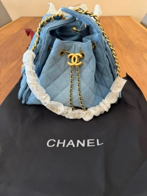 Blå quiltad handväska  - Säljer en snygg blå quiltad handväska i denim med guldfärgade kedjedetaljer och klassisk CC-logga framtill. Väskan har dragskostängning och en ficka på sidan med logga. Perfekt för dig som vill ha en trendig och exklusiv accessoar.