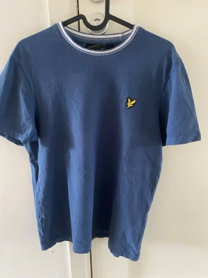 Blå t-shirt från Lyle & Scott - Snygg blå t-shirt från Lyle & Scott med klassisk rund halsringning och gul logga på bröstet. Tillverkad i mjuk bomull och har en enkel, stilren design som passar till det mesta. Perfekt för dig som gillar en clean och avslappnad look.