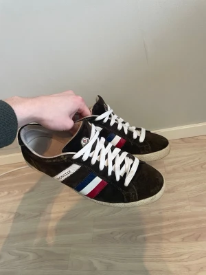 Moncler sneakers i mocka - Snygga Moncler sneakers i mörkbrun mocka med vita snören och klassiska Moncler-ränder. Säljer dessa för att jag inte passar i dom längre. Gör gärna av er vid minsta fråga. Kan sänka pris vid snabb affär