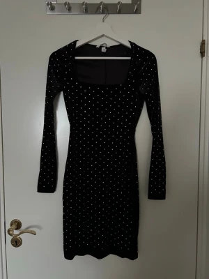 Svart sammetsklänning med silvriga stenar  - Snygg svart sammetsklänning med silvriga stenar och långa ärmar. Klänningen har en fyrkantig ringning och figurnära passform som ger en elegant siluett. 