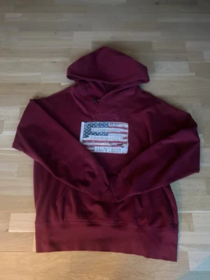 Vinröd hoodie från Ralph Lauren - Snygg vinröd hoodie från Ralph Lauren med stor amerikansk flagga och logga broderad på bröstet. Klassisk känguruficka framtill och huva med dragsko. Tillverkad i mjukt bomullsmaterial, perfekt för en avslappnad och cool stil.