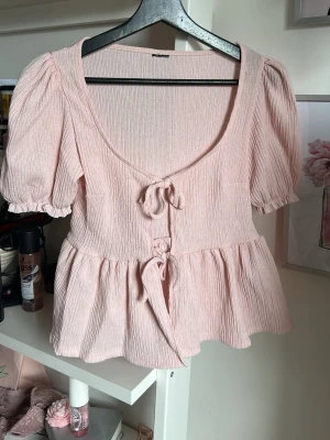 Ljusrosa blus med knyt och puffärm - Supersöt ljusrosa blus med puffärmar och två knyt framtill. Blusen har en djup rundringning och volang nedtill, vilket ger en romantisk vibe. Perfekt för dig som gillar feminina detaljer och en luftig känsla.
