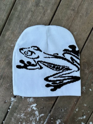 Vit mössa med svart grodmotiv - Säljer en vit mössa med ett stort svart grodmotiv på sidan. Mössan är slätstickad och har en klassisk, något längre passform. Perfekt för dig som vill sticka ut med en unik accessoar. 