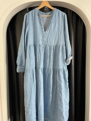 Ljusblå långklänning i linne - Långklänning i ljusblått linne med v-ringning och långa ärmar. Klänningen har en avslappnad passform och faller snyggt i flera lager. Perfekt för en chill sommardag eller när du vill vara bekväm men ändå stilren.