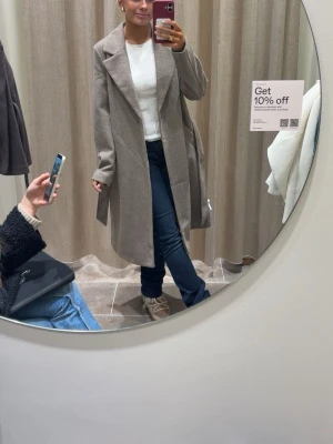 Beige kappa med skärp och bred krage - Beige kappa med bred krage och knytskärp i midjan. Lång modell med raka ärmar  Perfekt för dig som gillar en klassisk och clean look. Materialet ser ut att vara ullblandning för extra värme.