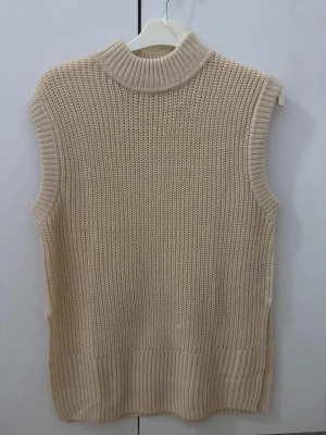 Beige stickad väst med polokrage - Säljer en beige stickad väst med hög polokrage och bred ribbad kant nedtill. Västen är ärmlös och har en avslappnad passform, perfekt att styla över en skjorta eller t-shirt. Materialet är mjukt och grovstickat för en trendig look.