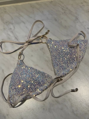 Glittrig ljuslila bikiniöverdel - Supersnygg trekantsbikini med ljuslila bas och massor av glittrande paljetter. Bikinin har tunna knytband och guldfärgade ringdetaljer. Perfekt för dig som vill sticka ut på stranden eller poolpartyt.