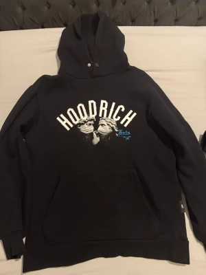 Svart hoodie från Hoodrich med tryck - Svart hoodie från Hoodrich med stor huva och känguruficka. Framsidan har ett vitt tryck med texten 'HOODRICH' och konstmotiv, samt blå detaljer. Baksidan har ett stort konstnärligt tryck med text och grafiska element i vitt och blått. Perfekt för en streetwear-look.