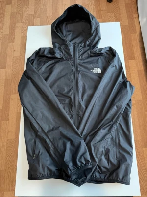 vindjacka The North Face - Snygg svart vindjacka från The North Face med huva och dragkedja framtill. Jackan har två sidofickor med dragkedja och logga på bröstet samt ryggen. Tillverkad i lätt och tåligt material som står emot vind. Perfekt för blåsiga dagar.