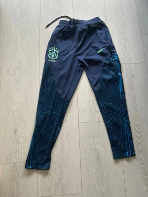 Nike Brasil träningsbyxor blå - Snygga träningsbyxor från Nike med Brasil-logga och Nike Swoosh i grönt. Byxorna är mörkblå med grafiskt mönster i ljusblått på benen, har resår i midjan och dragsko för bästa passform. Perfekta för fotbollsträning eller gymmet.
