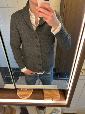 Grå kofta med sjalkrage från Esprit - Säljer en grå stickad kofta från Esprit med sjalkrage och snyggt mönster i tyget. Koftan har bruna knappar framtill, två stora fickor och ribbade muddar vid ärmslut och nederkant. Perfekt att slänga på över en skjorta för en chill men stilren look.