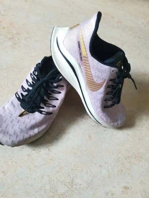 Nike Zoom Pegasus lila sneakers - Säljer ett par Nike Zoom sneakers  med svarta skosnören och detaljer i guld och svart. Skorna har en vit och svart sula med bra dämpning och den klassiska Nike-loggan på sidan. Perfekta för dig som gillar sportig och stilren design.