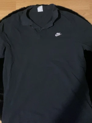 Svart pikétröja från Nike - Klassisk svart pikétröja från Nike med vit logga broderad på bröstet. Tröjan har krage och knappar framtill, perfekt för en sportig och clean look. Tillverkad i mjuk bomull som känns skön mot huden.