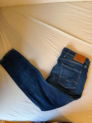 Mörkblå jeans från Replay - Säljer ett par mörkblå jeans från Replay med klassisk femficksmodell och raka ben. Jeansen har snygga slitningar och kontrastsömmar samt en läderpatch bak i midjan. Tillverkade i bomull för en skön och slitstark känsla.