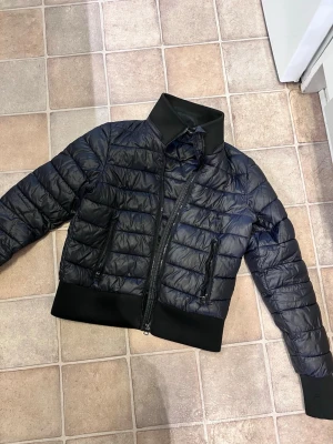 Svart vårjacka med hög krage - Snygg svart jacka med hög krage och breda muddar i ärmslut och nederkant. Jackan har två dragkedjeförsedda fickor framtill och quiltad design. Perfekt för kalla dagar och ger en modern look.