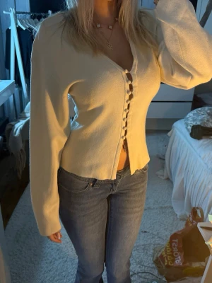 Krämvit kofta med knappar - Säljer en krämvit kofta med figurnära passform och dekorativa knappar framtill. Koftan har långa ärmar och en lätt v-ringad halsringning. Perfekt att styla med jeans eller kjol för en trendig look.