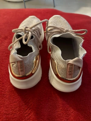 Beige sneakers från Adidas med guld - Stilrena sneakers från Adidas i beige stickad textil med vita sulor och snygga gulddetaljer vid hälen. Skorna har elastiska snören och en modern, vågig kant runt öppningen. Perfekta för dig som gillar sportig och trendig stil.