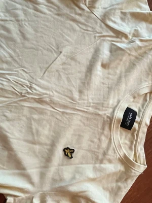 Gul t-shirt från Lyle & Scott - Snygg gul t-shirt från Lyle & Scott med klassisk rund hals och det ikoniska gulsvarta fågelmärket broderat på bröstet. Tillverkad i mjuk bomull som känns skön mot huden. Perfekt till jeans eller shorts för en clean och enkel stil.