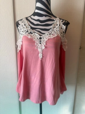 Rosa linnetopp med spetsdetaljer - Supersöt rosa linnetopp med vita spetsdetaljer runt ringningen och axelbanden. Toppen har en v-ringad design framtill och är ärmlös med en lös passform. Perfekt för dig som gillar romantiska och feminina plagg.